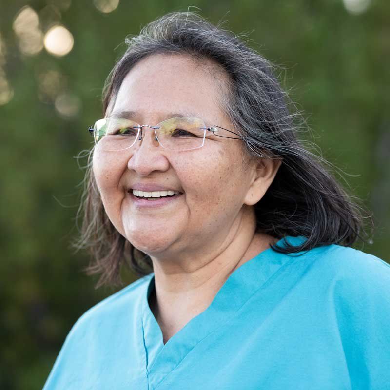 Adriann Begay, late middle aged wearing rimless glasses and straight dark hair, smiles past the camera in a shoulders-up portrait.