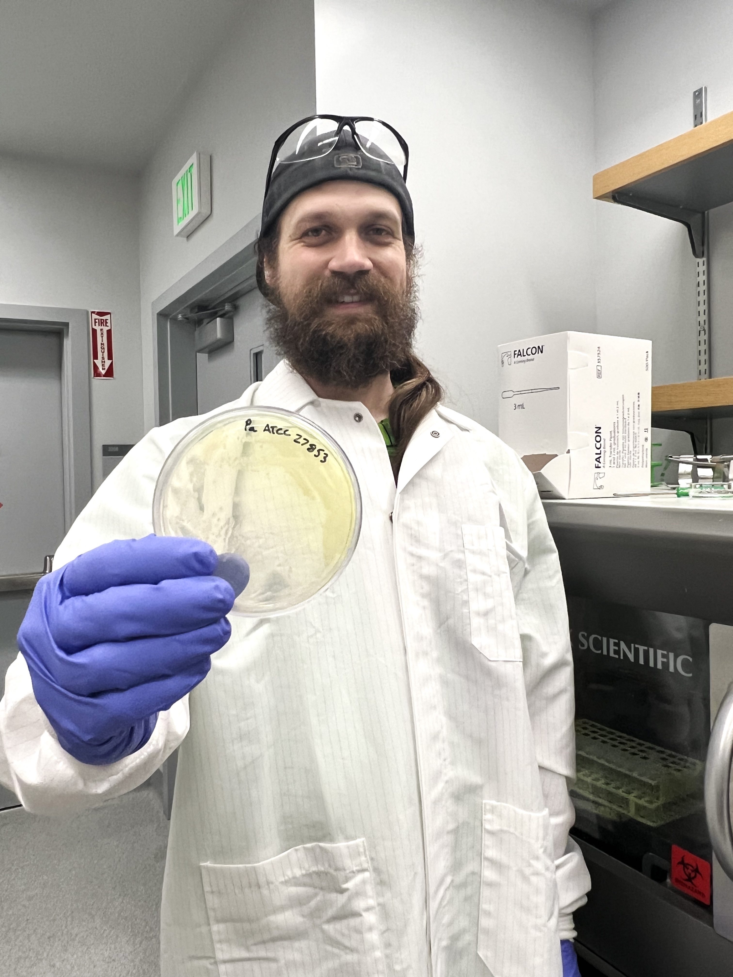 Man with big bushy beard, backwards ballcap, safety glasses atop his head, wearing white lab coat and purple rubber gloves, holds up a cell culture plate, smiling for camera in a lab