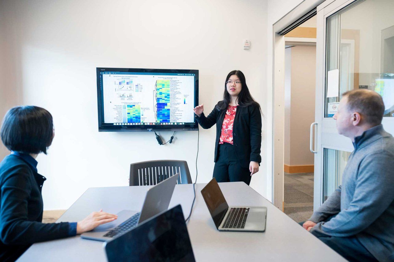 A young woman of Asian descent explains an image on a large computer monitor to two people seated at a table