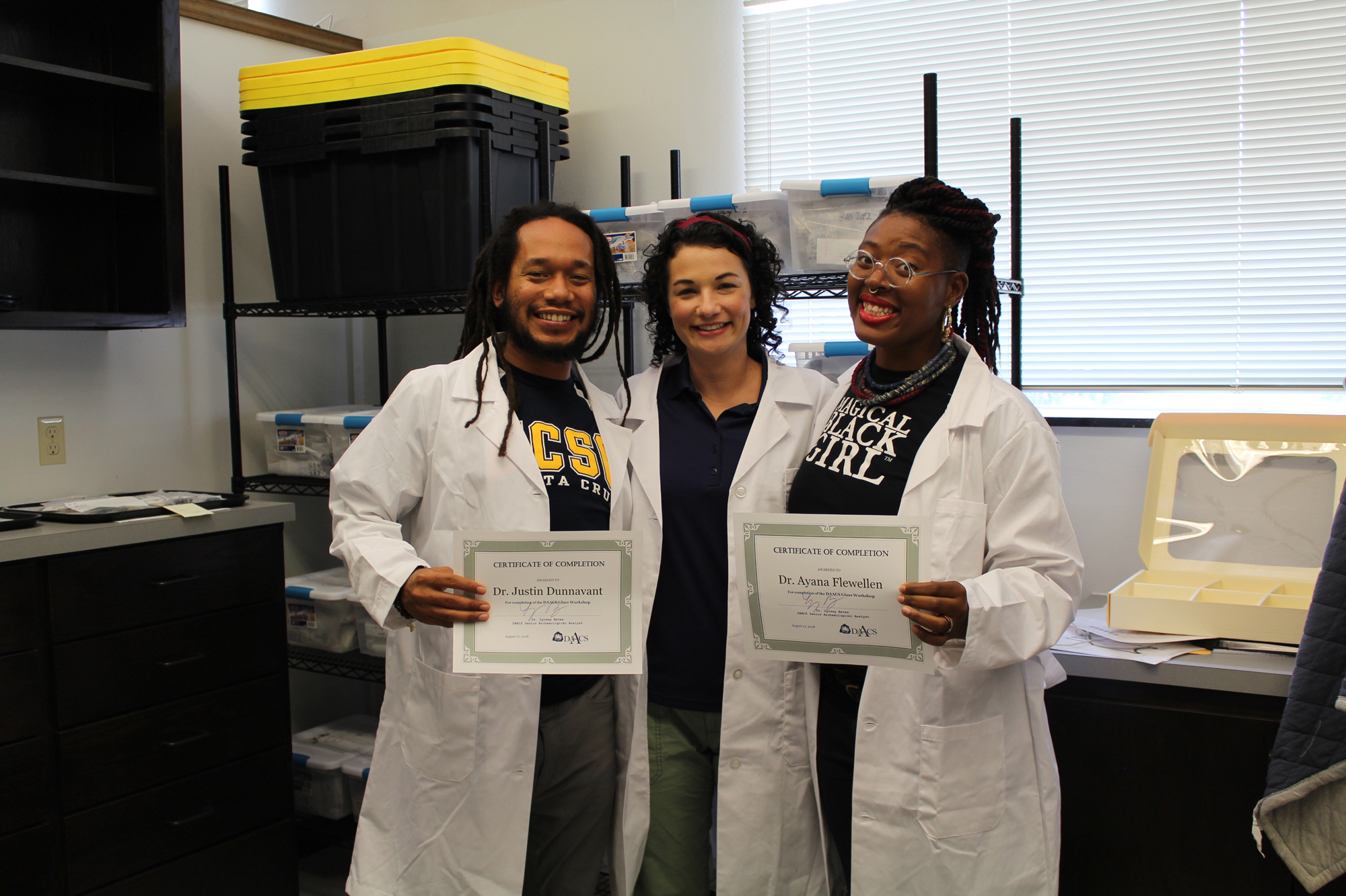 A young African American male, a white woman and an African American nonbinary person hold up certificates while wearing lab coats and smiling