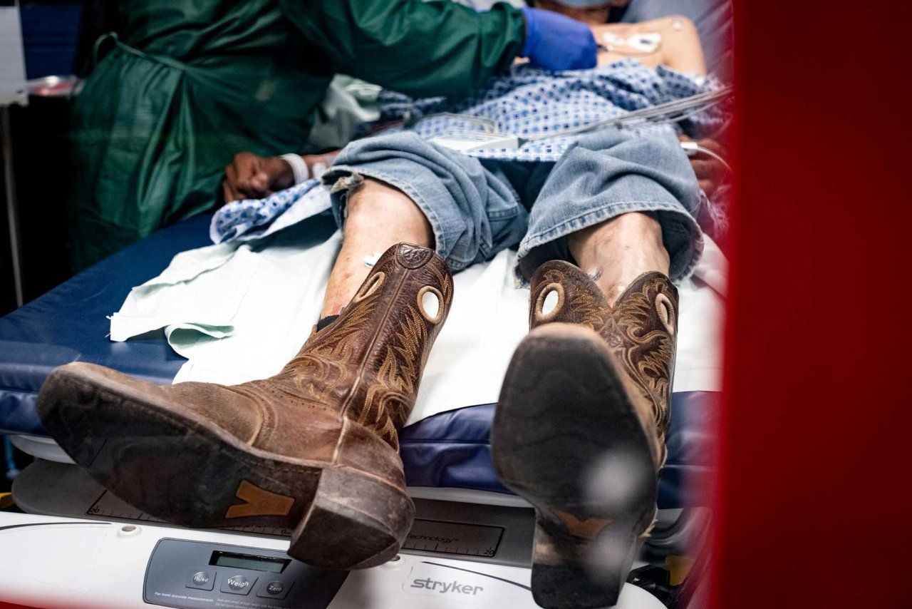 An elderly person lying in a hospital bed, being tended to by a provider wearing a green gown and blue gloves. The patient's jeans are pulled up above their knees, showing scuffed cowboy boots hanging off the edge of the bed.