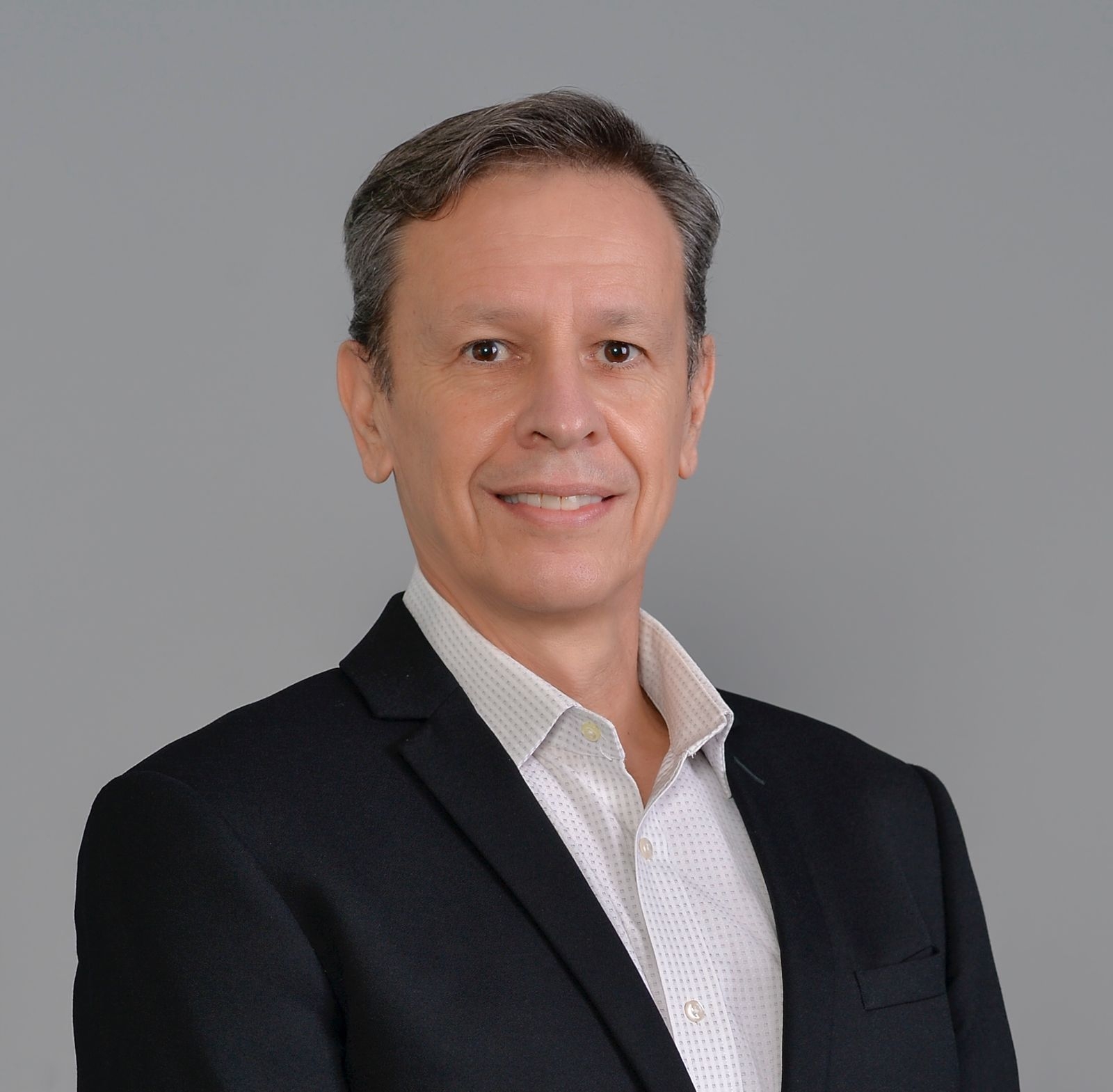 Middle aged man with brown hair smiles for a professional headshot wearing white button up shirt and black blazer against gray background