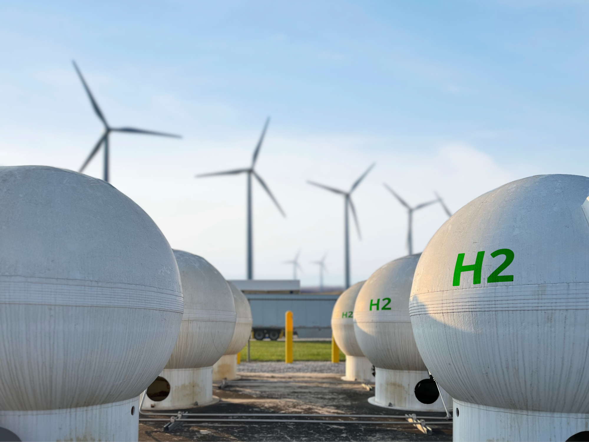 Two rows of spherical white tanks with H2 printed on them in green, with four wind turbines in the background, against a blue sky