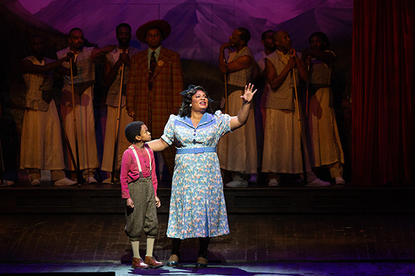 A young boy portraying Malcom X stands with a grown woman gesturing to the audience on the stage, with more adults in low lighting in the background