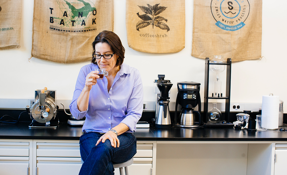 A white woman drinks coffee on a stool in a coffee shop