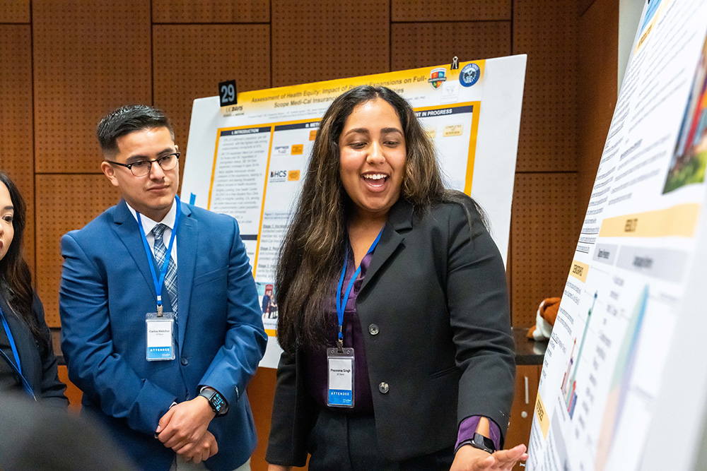 A young woman in a black shirt and conference lanyard presents a poster summarizing research to a group including a young man in glasses and a blue suit.