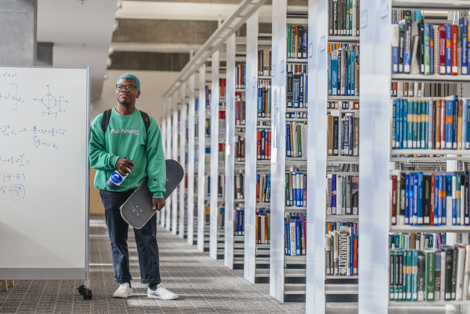 Young man wearing green sweatshirt carrying a skateboard leans against a white board next to library shelves