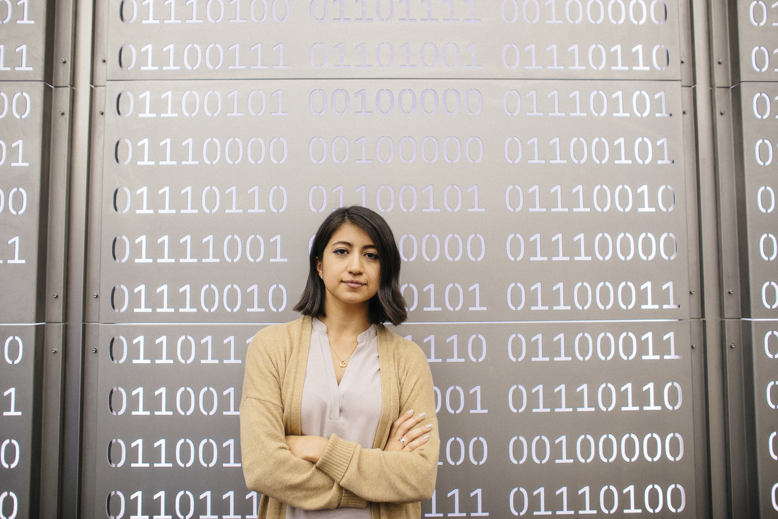 Woman crosses arms over chest and looks at camera, against a metal wall with punched out 0s and 1s, meant to represent computer code