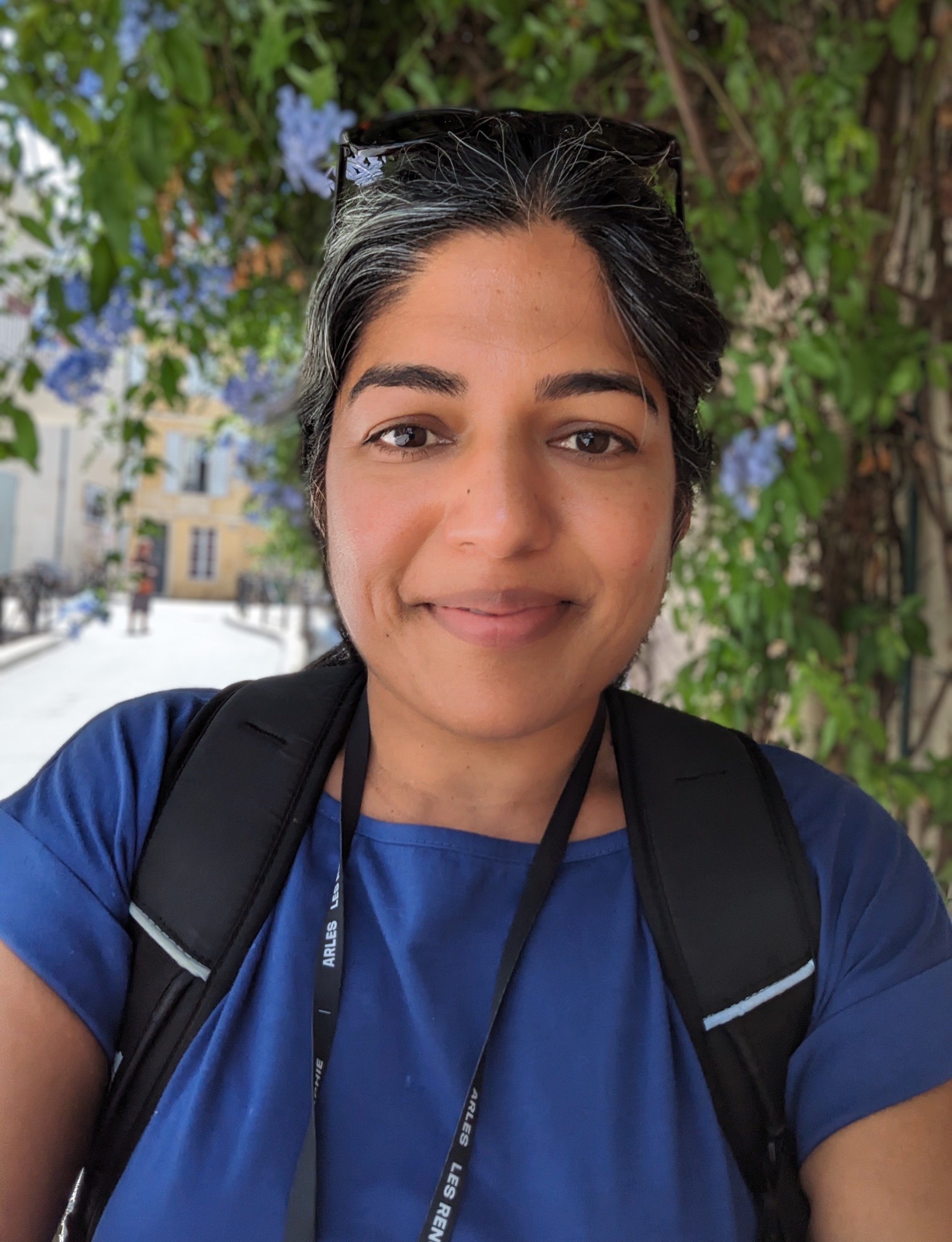 Jyoti Mishra, in blue scrubs, backpack, and lanyard, smiles for a selfie with a tree in the background