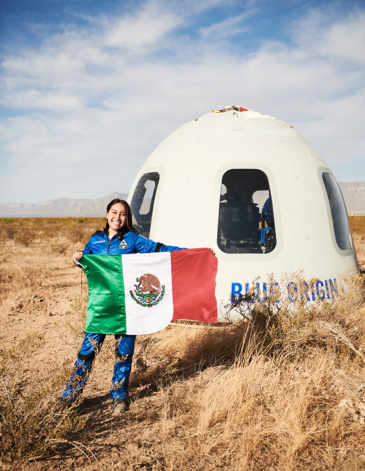 Katya Echazaretta wearing a blue jumpsuit holding a Mexican flag standing in front of a space capsule with "Blue Origin" written on the side, parked in a field of brown grass