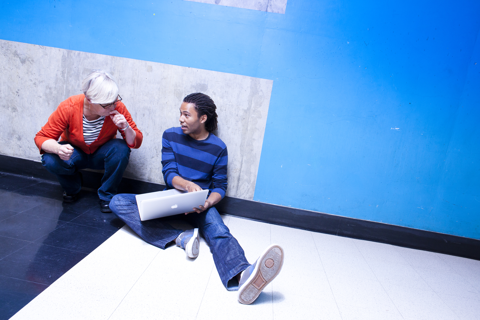 A female professor crouches next to a male student, who's sitting with his legs stretched out in a hallway with a blue wall, pointing to something on a Mac laptop screen