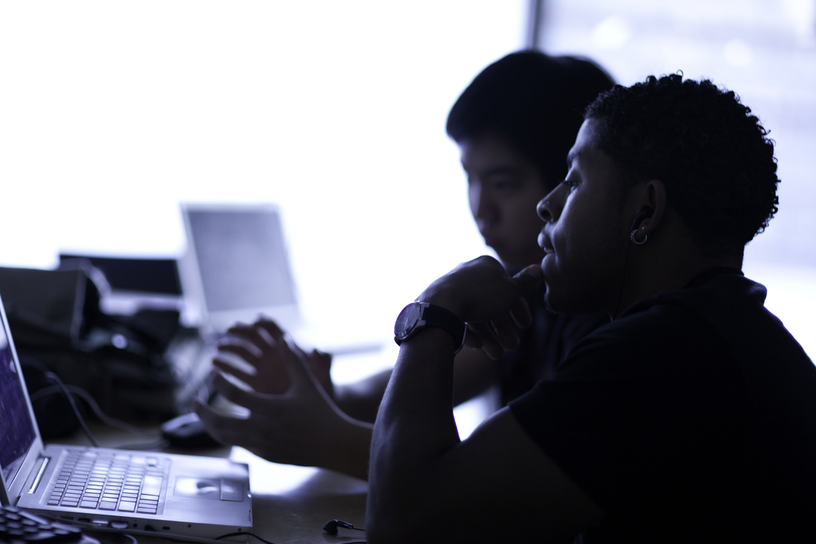 Two male college students type on laptops. Their profiles are visible but they're mostly backlit.