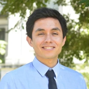 Francisco Castro smiles for a shoulders-up portrait wearing a blue shirt and tie against a backdrop of greenery