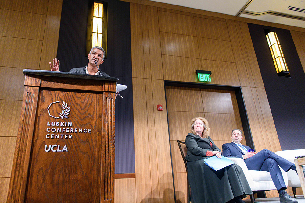 Ramesh Srinivasan gesticulates at a podium branded with UCLA Luskin Center. UC Provost Katherine Newman and MIT Professor Daron Acemoglu sit on stage next to the podium.