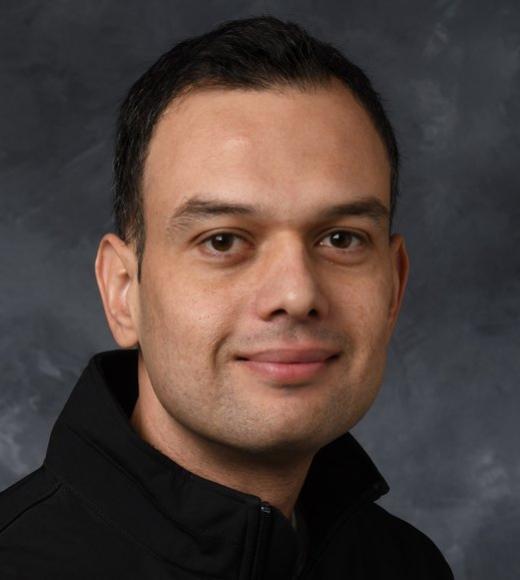 Zubair Shafiq, wearing a dark shirt, smiles for a shoulders-up portrait against a gray background