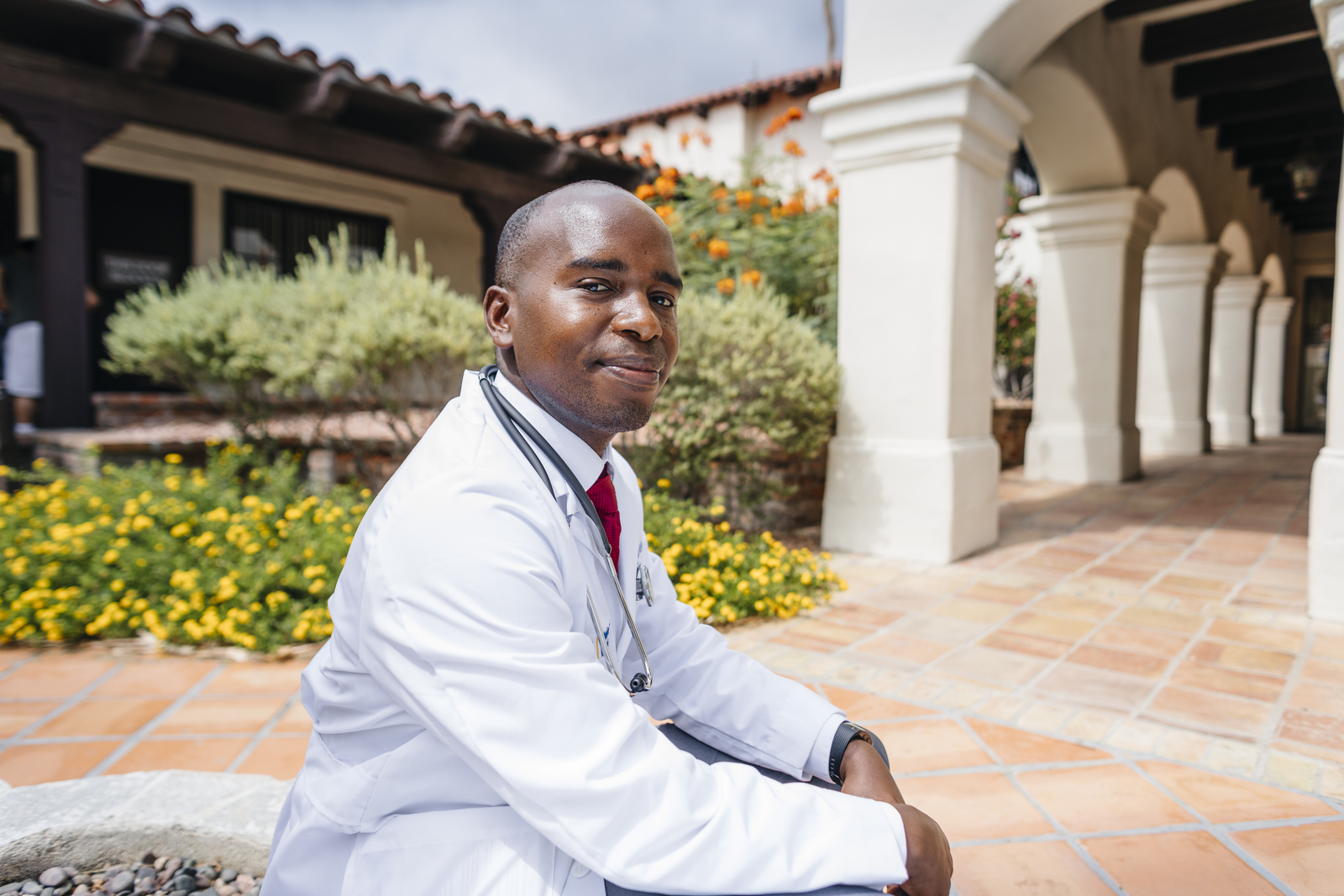 A male doctor in a white coat sits in a breezeway with greenery and arched walls