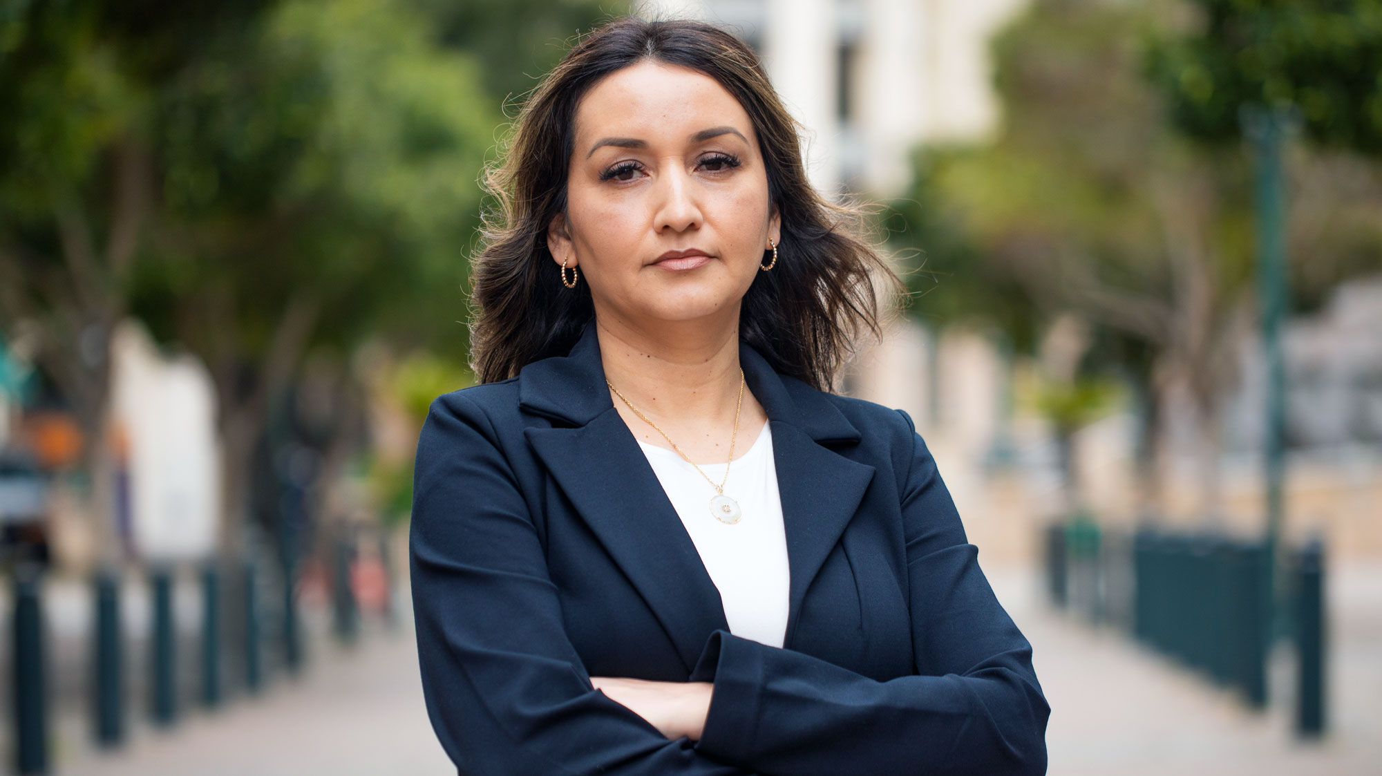 Daniela Medina, in a blazer and white shirt, folds arms over chest and looks at the camera, outside