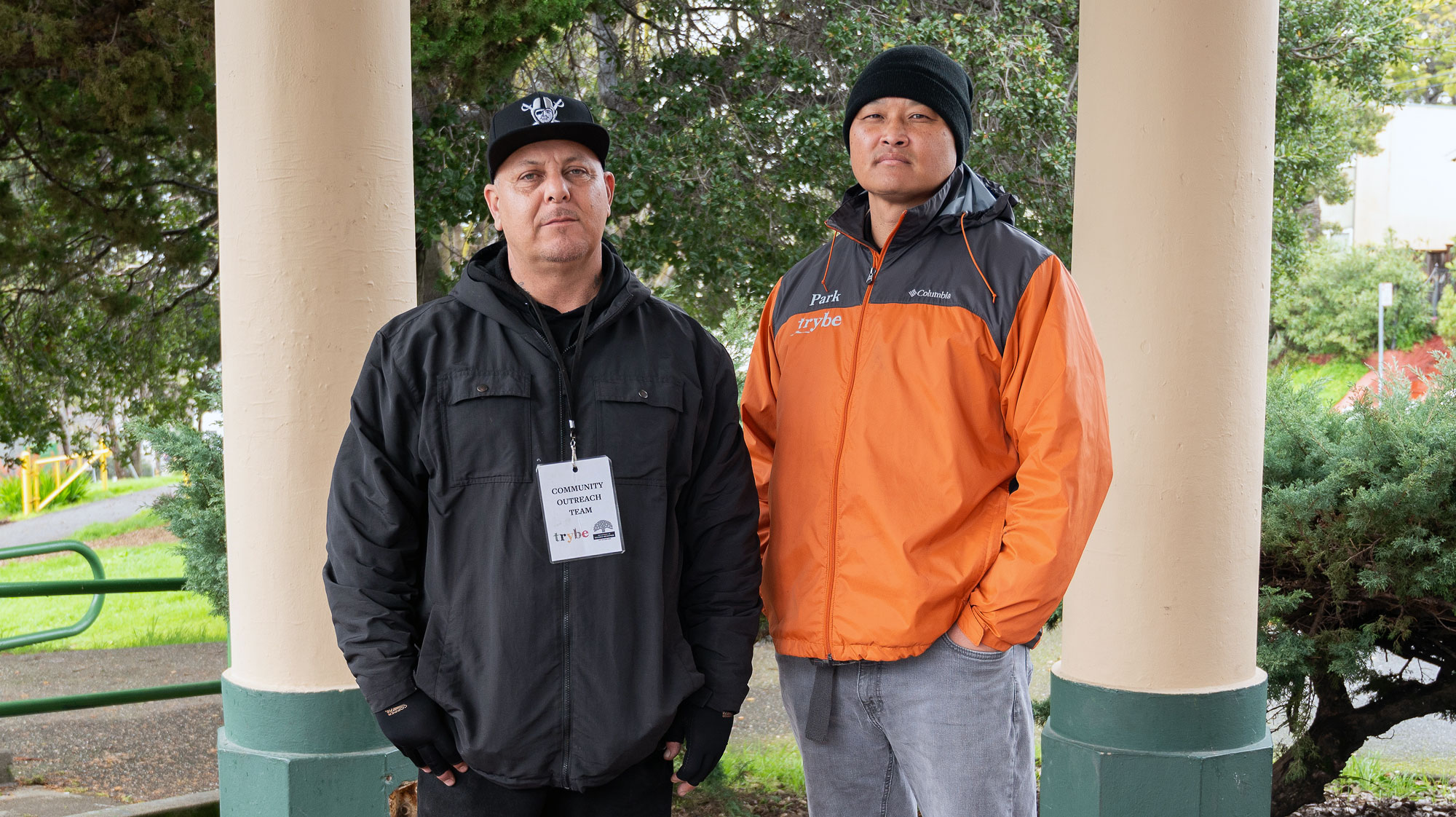 Hector Cruz and Andrew Park look at camera wearing cool weather clothing, standing under a pergola at a park