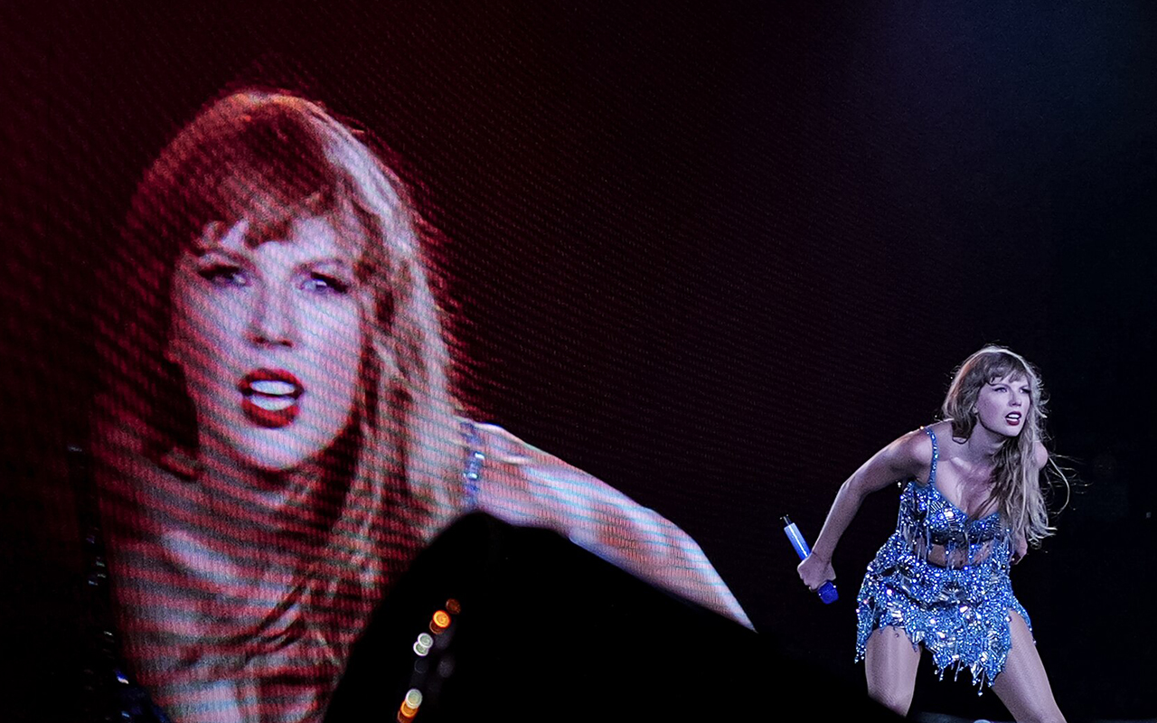 Taylor Swift performs in a sequined dress in front of an image of herself on a giant screen