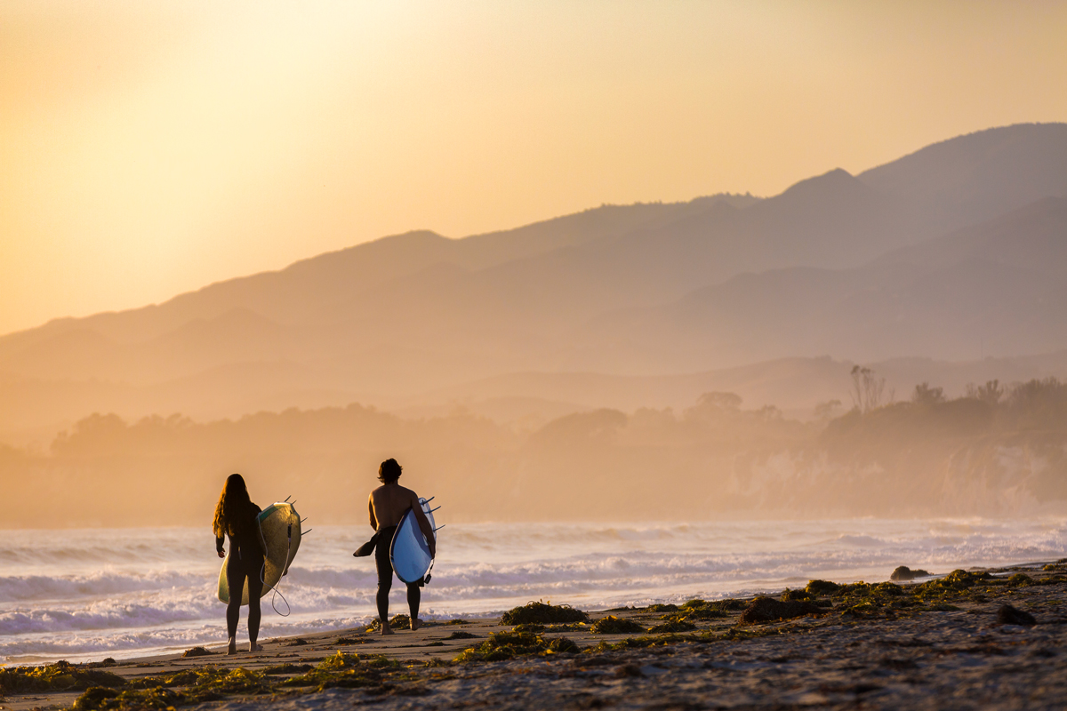 two surfers walk along a beach at sunset with mountains in the background