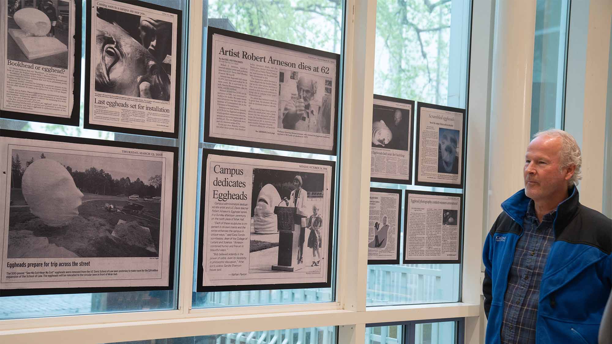 A bald man in a jacket (same man as the hero/first photo at the top of the article) looks at a collection of news clippings on the wall