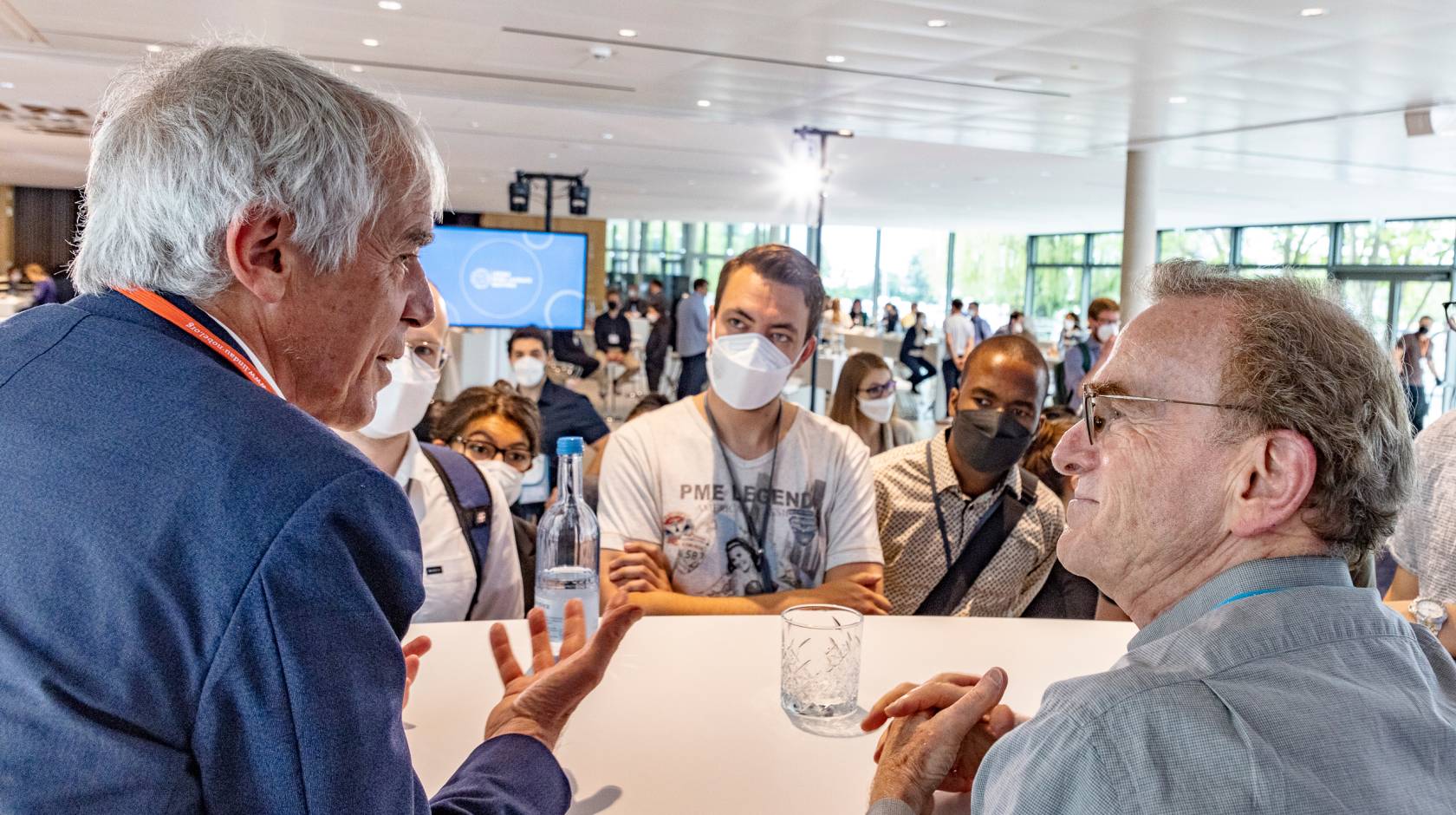 A photo of two older Nobelists talking with a group of young people in masks in front of them listening