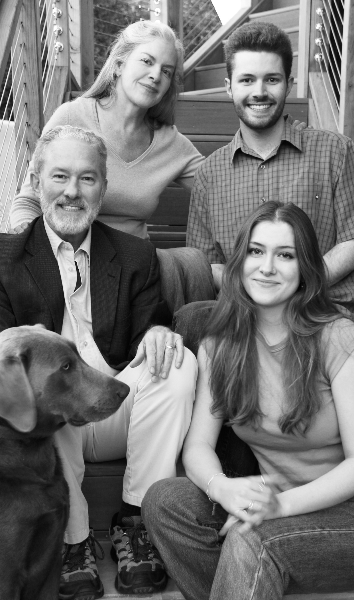 A black and white photo of Rich Lyons and his family seated on stairs with his daughter seated next to him; wife seated above; son seated next to her; and Weimaraner dog poking his head in 