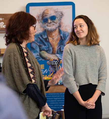 A woman with glasses faces a woman who is smiling in front of a painting of a bald bearded man with blue sunglasses