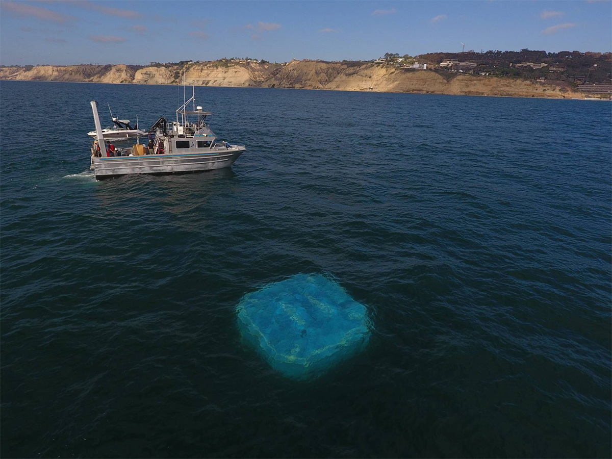 Blue, car-sized platform floats just below surface of placid ocean, with a metal fishing boat monitoring nearby
