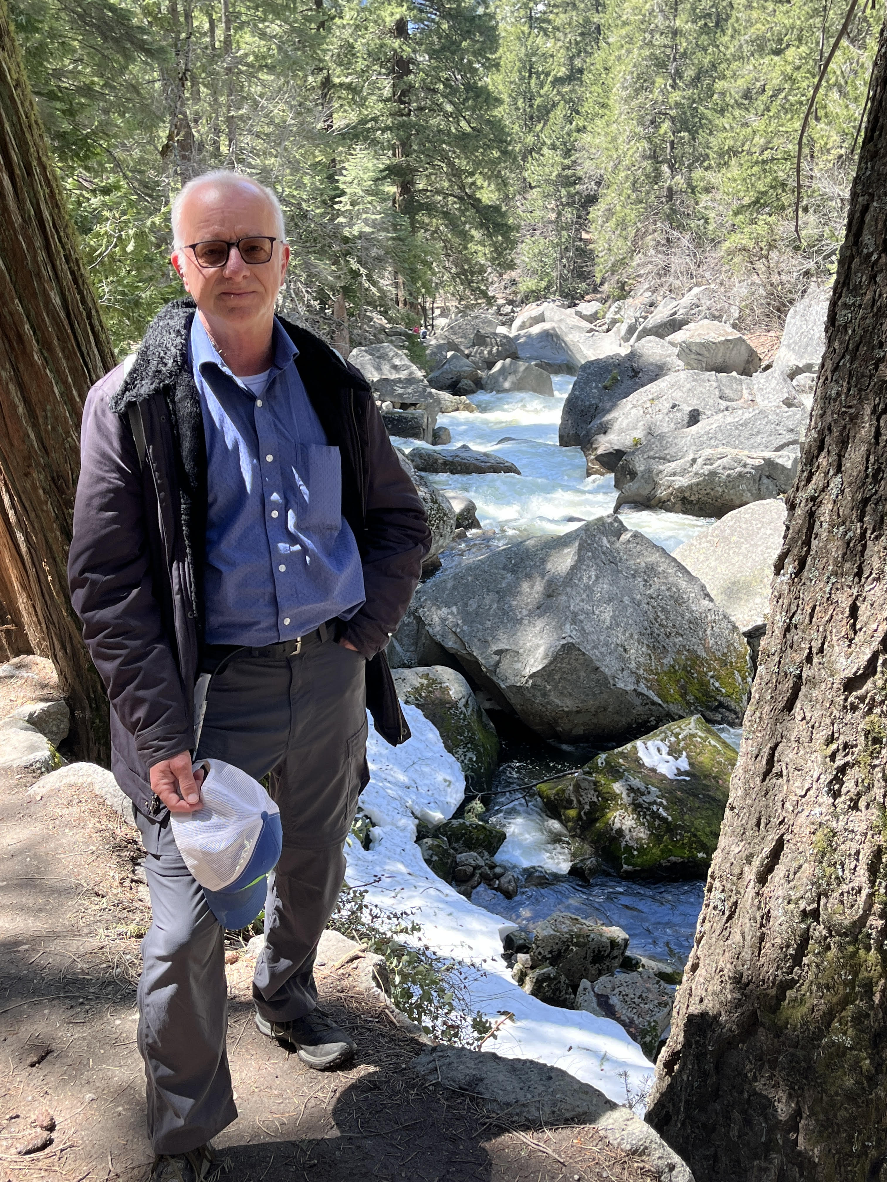Older man in a jacket, holding a hat, next to a creek in the Sierras
