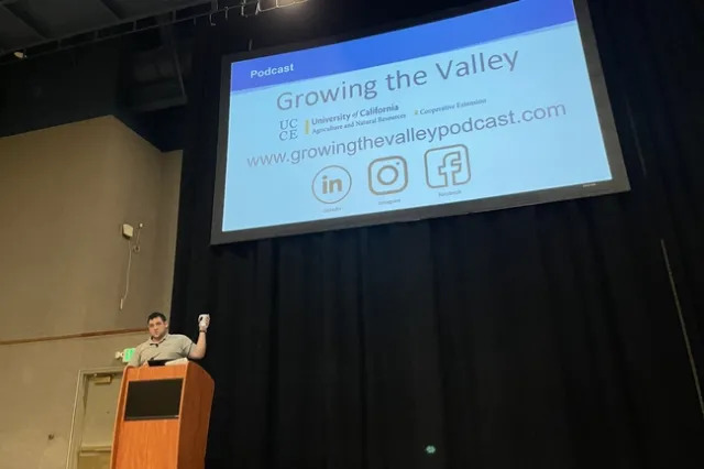 A man at a podium gestures at a large projection screen behind him that says "Growing the Valley"