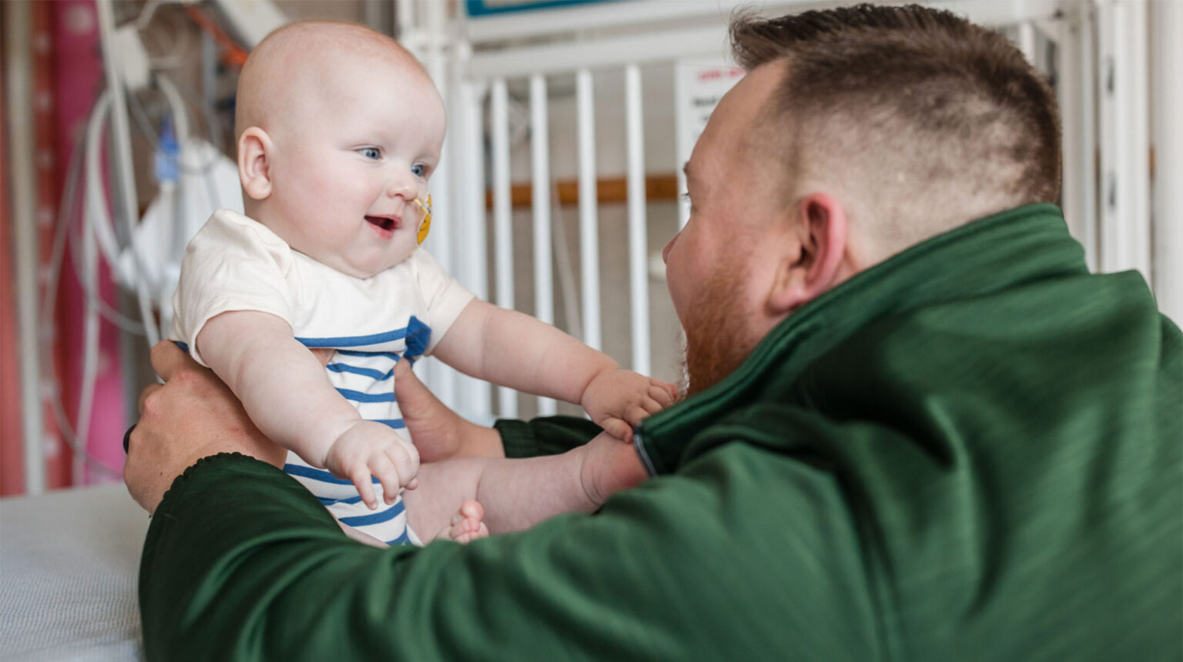 A baby with a tube in his nose smiles at his dad, who's holding him up and smiling back.