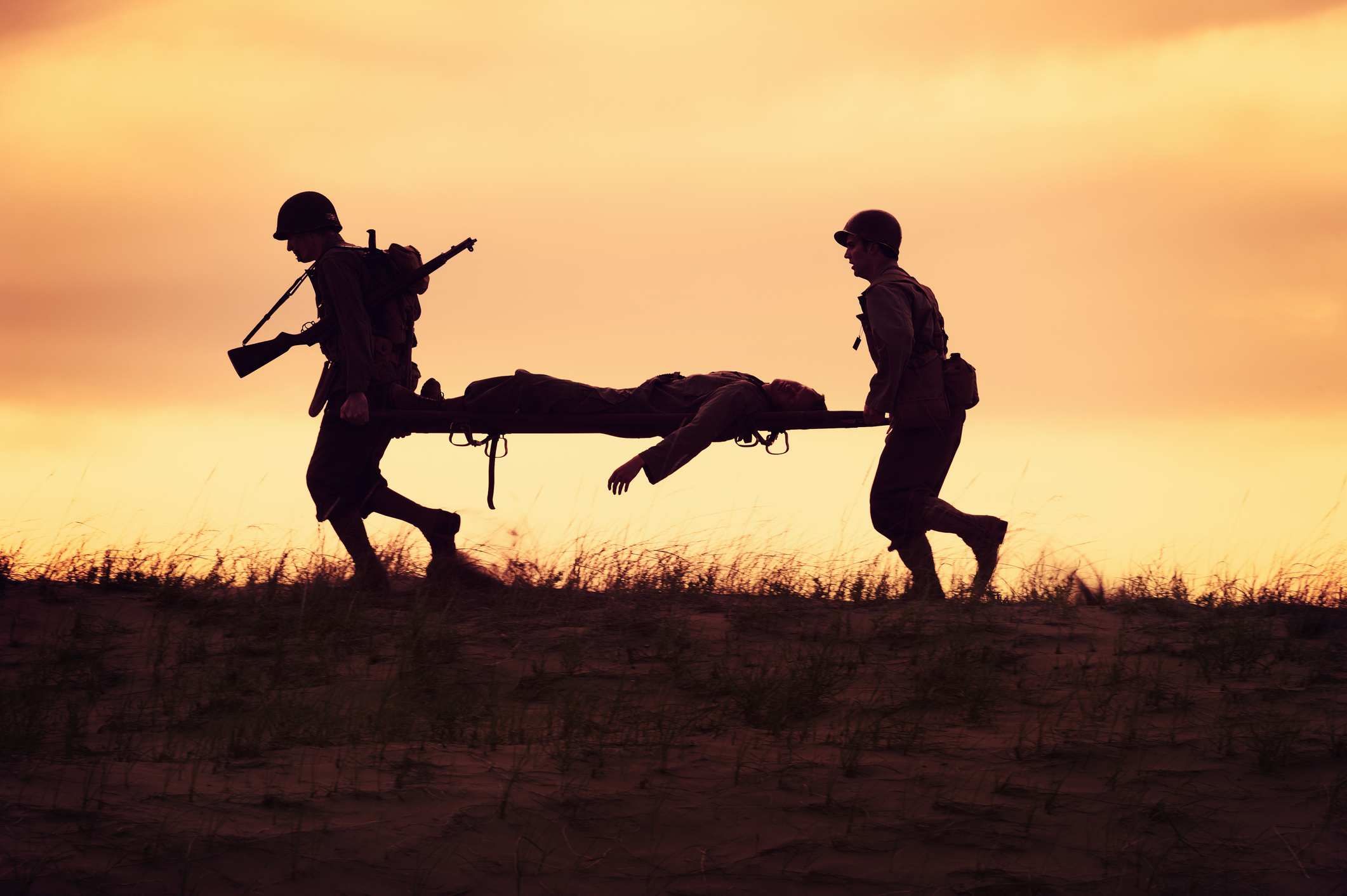 Two soldiers in silhouette carry a stretcher with another soldier on it