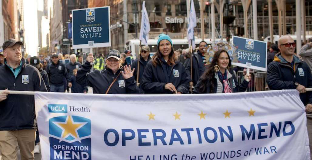People in blue uniforms march with an Operation Mend banner in front of them, one holds a sign that says Saved My Life