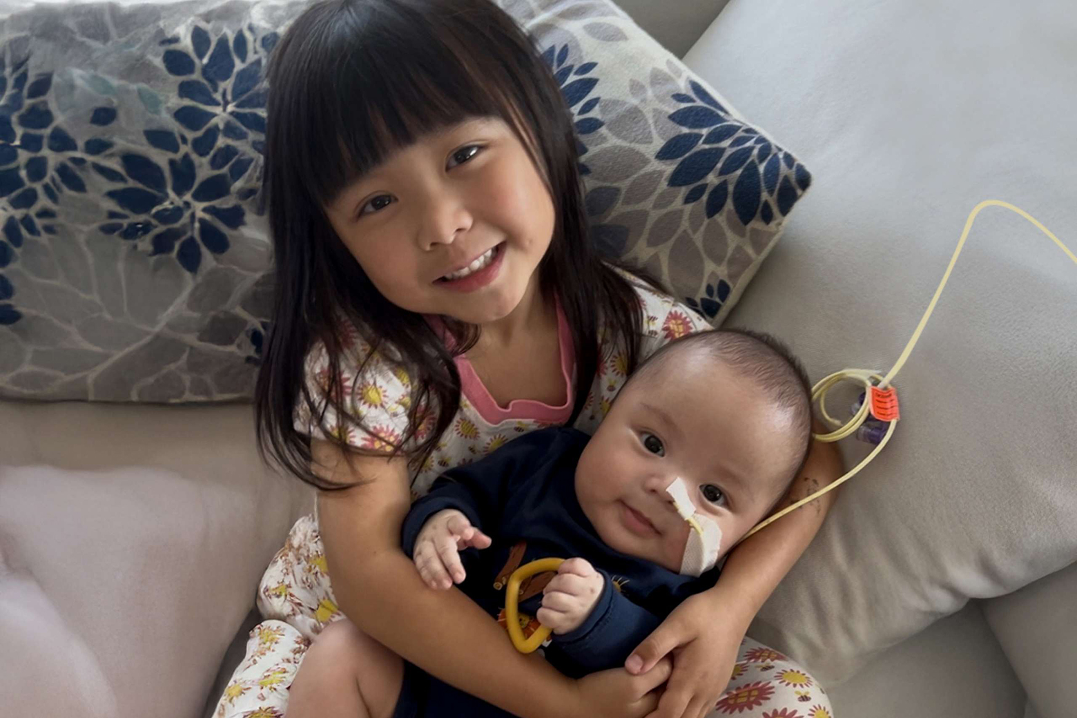 A kid sister holds her baby brother on her lap. The baby has a nasogastric tube.