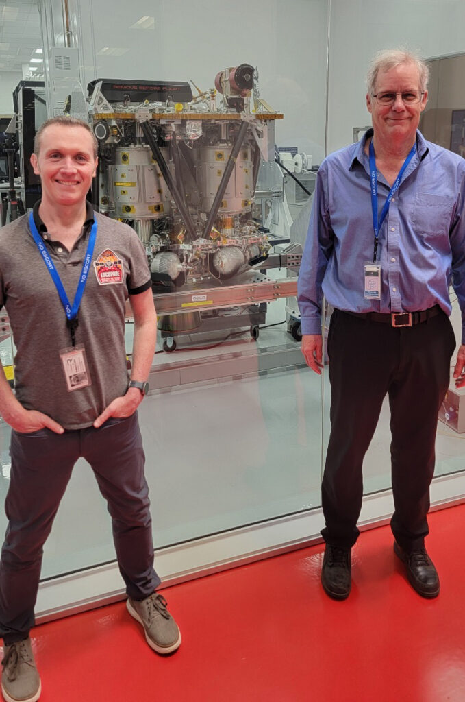 Two men stand on a red carpet in front of a glass wall looking into a room containing a large space-looking machine