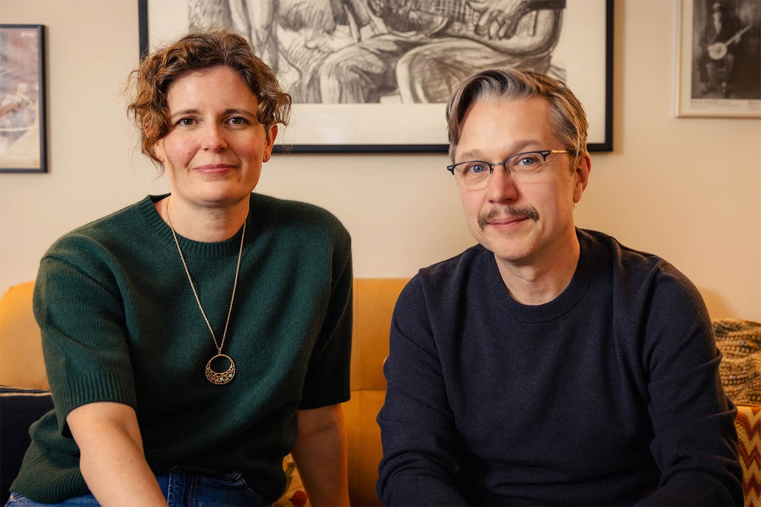 A man and a woman sit side-by-side on a couch. Illustrations of musicians hang on the wall behind them.