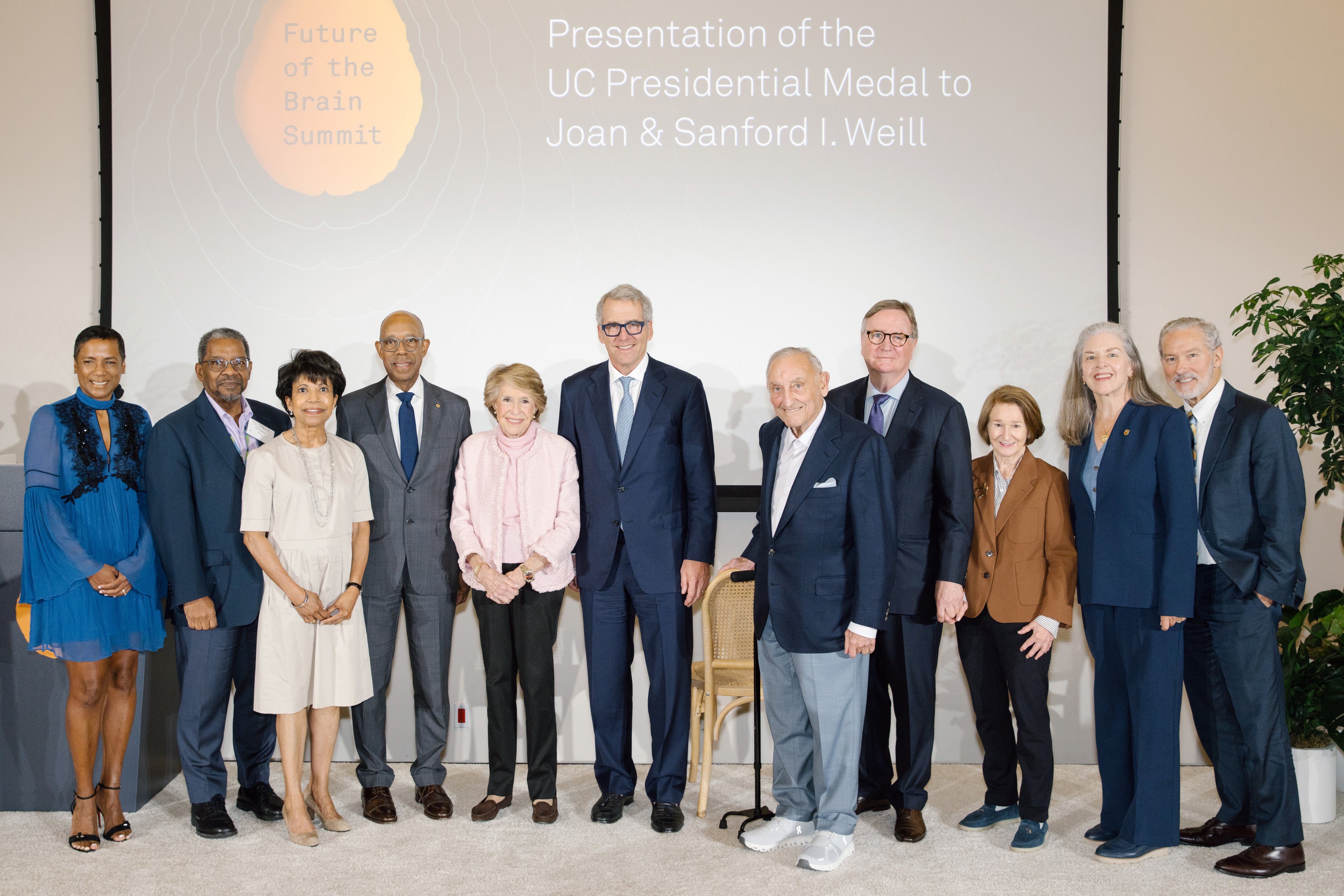 A group of about a dozen people in formal clothes pose on a stage in front of a projector screen