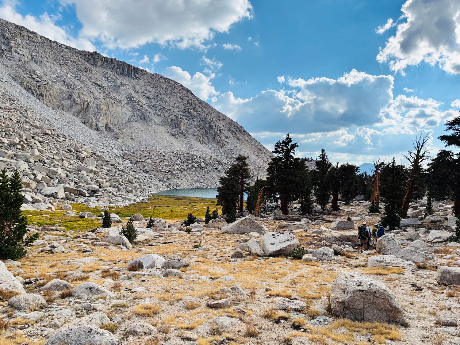 Wide-angle image of 2 people hiking with big backpacks through a dry meadow at the base of a tall mountain, towards a lake
