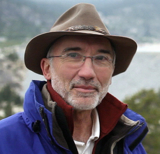 A man in a fedora-like hat with a beard photographed outside in a mountain range