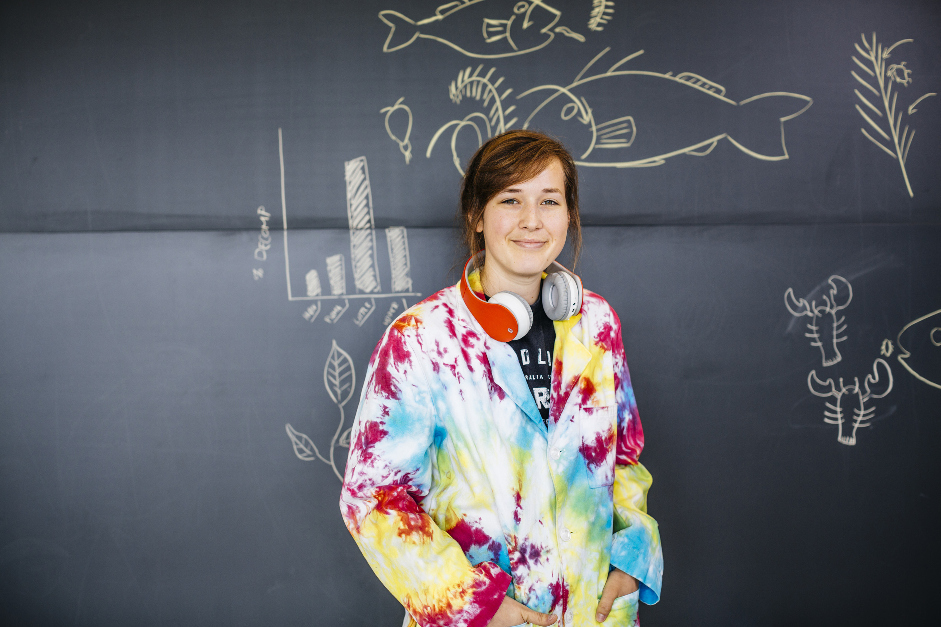 A young woman wearing headphones and a tie-dyed hoodie stands in front of a chalkboard with various drawings on it, including a few fish 