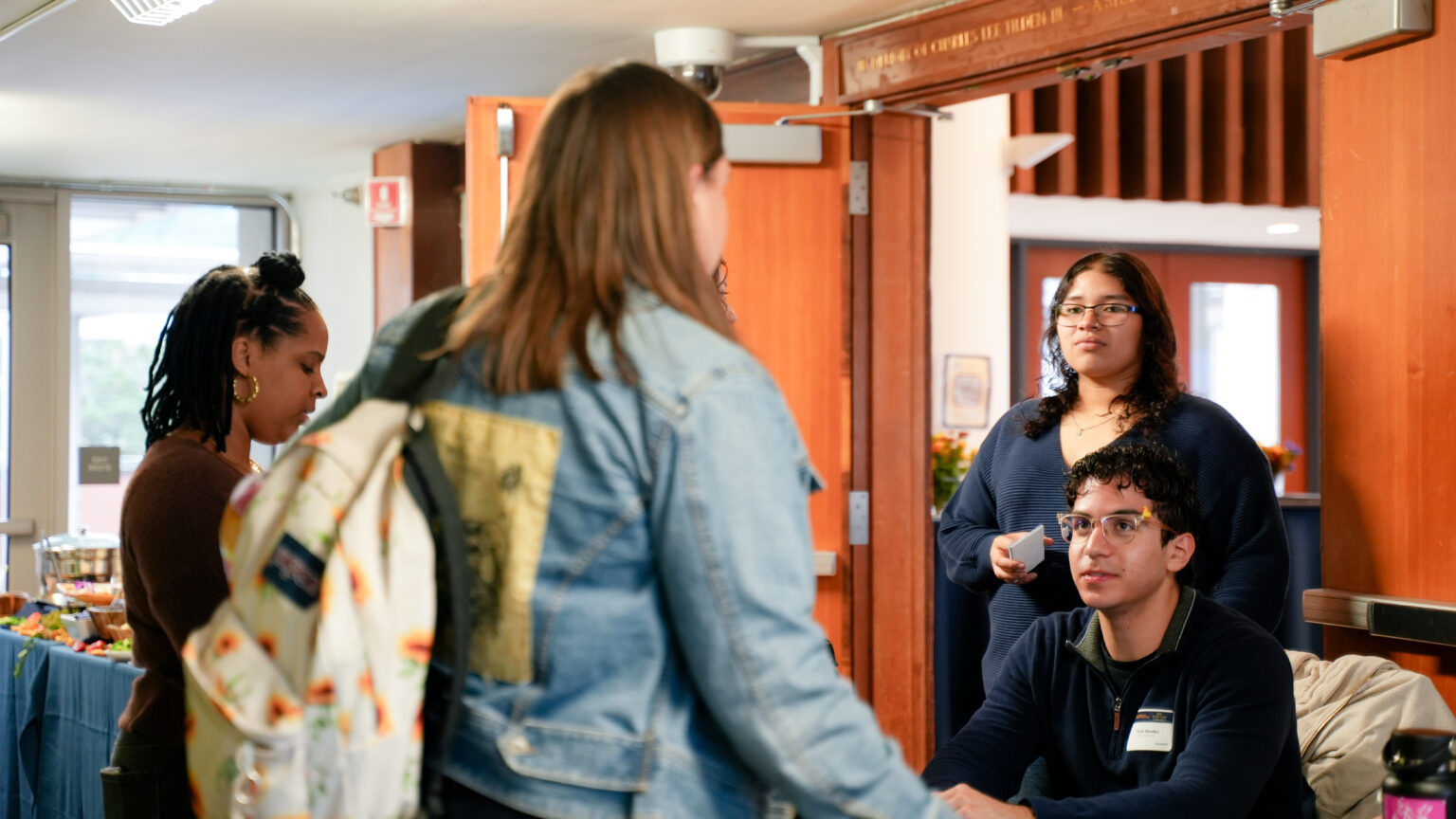 students of varying ethnicities talk at an event. One sits behind a table wearing a name tag.