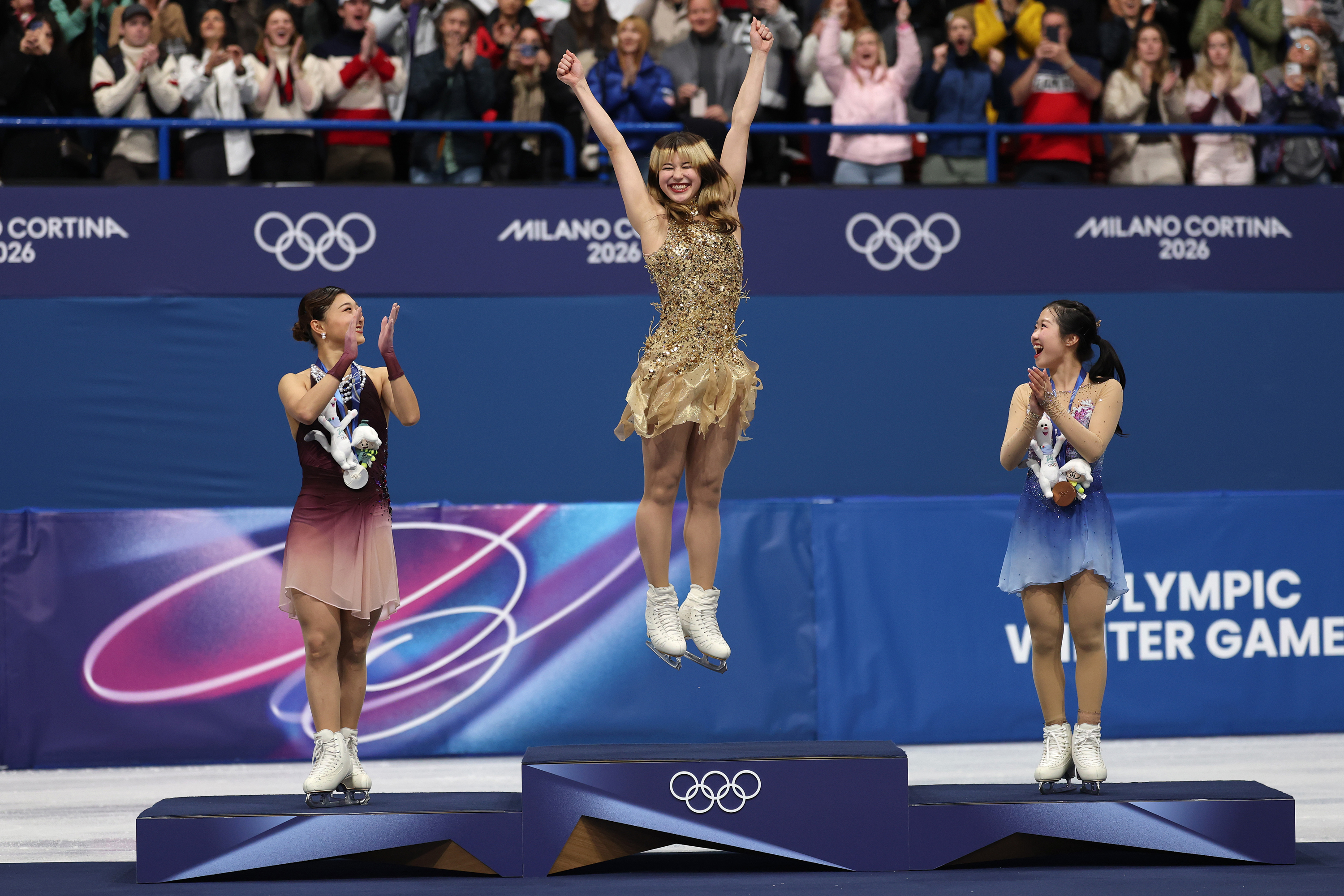 Three women skaters on an Olympics medal podium on ice; Alysa Liu, gold medalist, jumps in the air