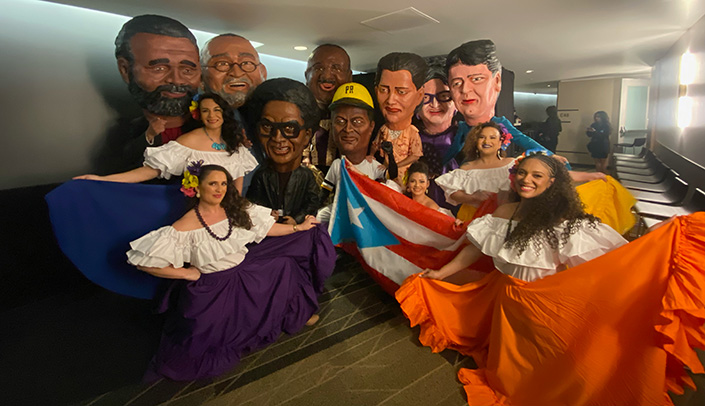 Several dancers display their costumes next to large decorative heads