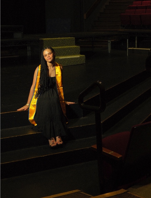 A woman in a graduation stole sits on a stage