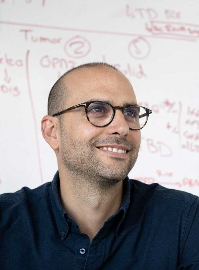 A man with glasses smiles, whiteboard behind him