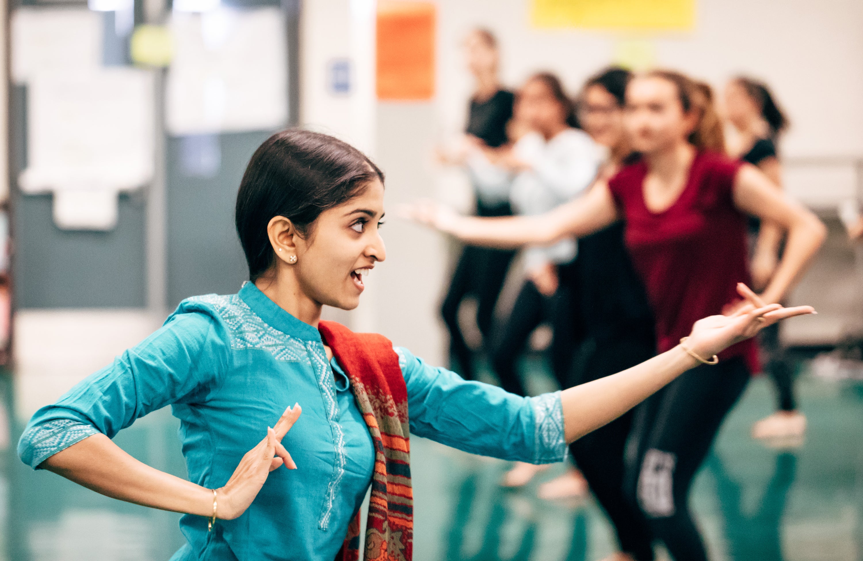A woman in blue demonstrates classical Indian dance techniques as students follow her in the background