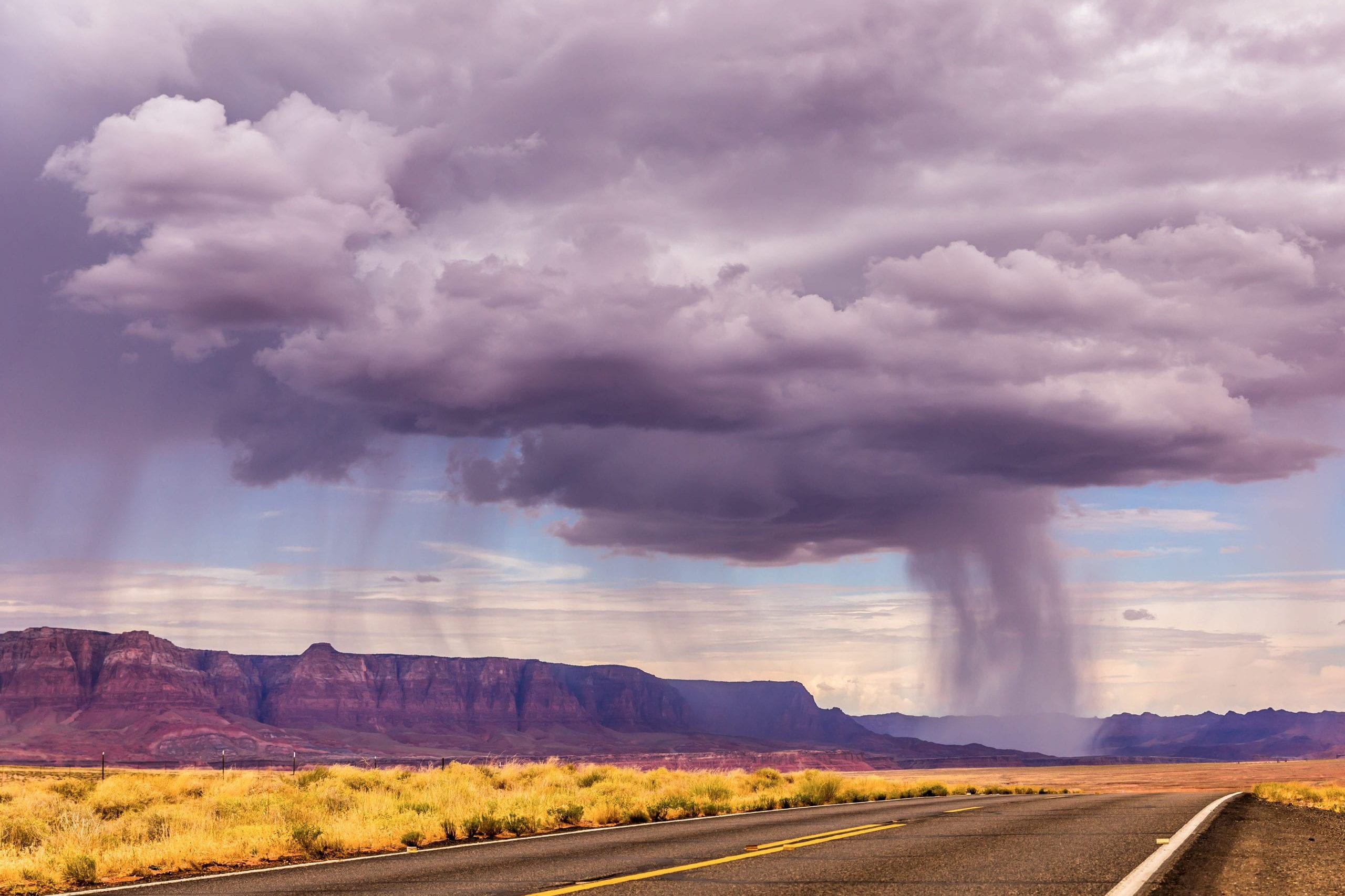 A desert landscape with a stretch of highway in the foreground and hills in the background, under a giant thundercloud with rain streaming down in the distance