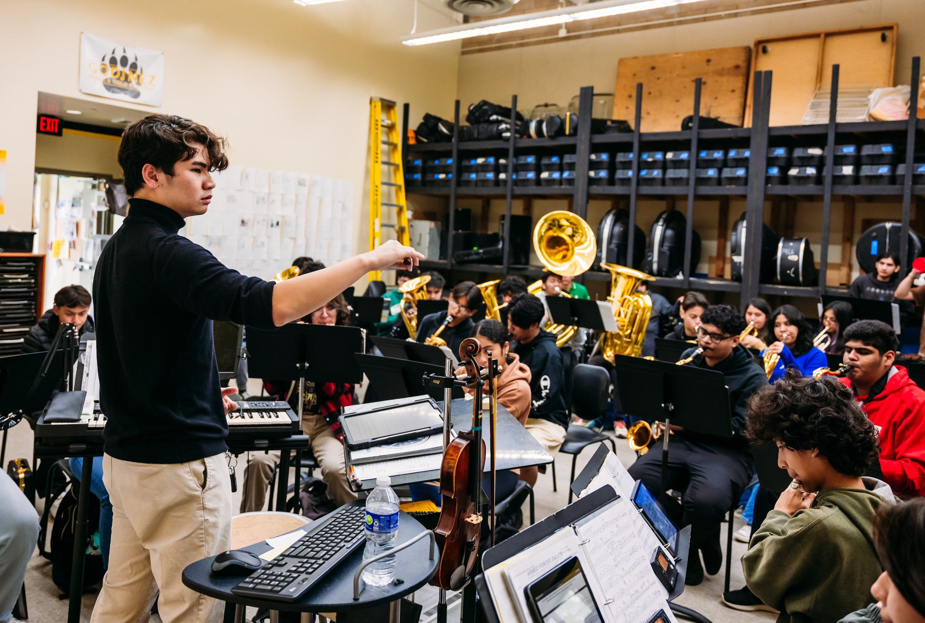 A young man conducts a high school student band in a rehearsal room