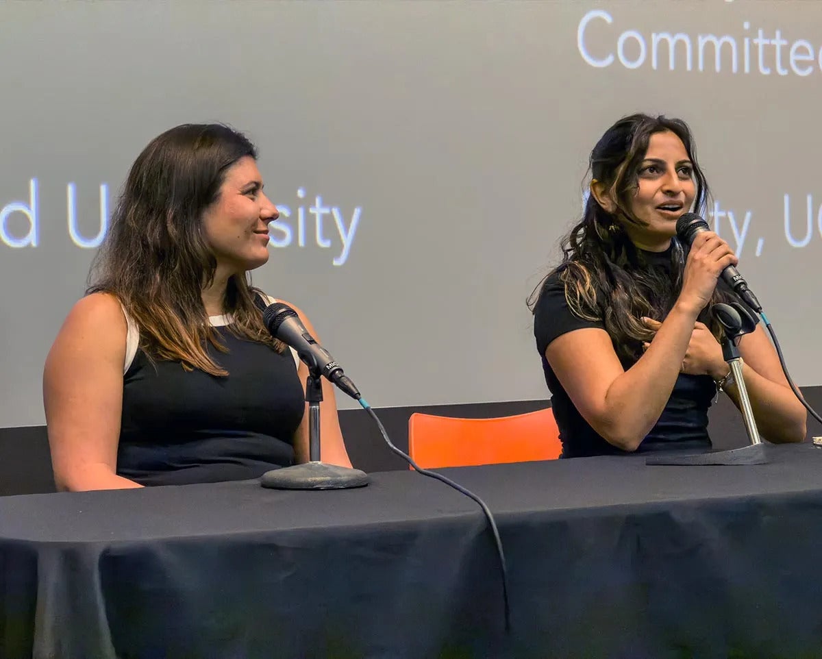 Two young women sit at a table with microphones, one is talking